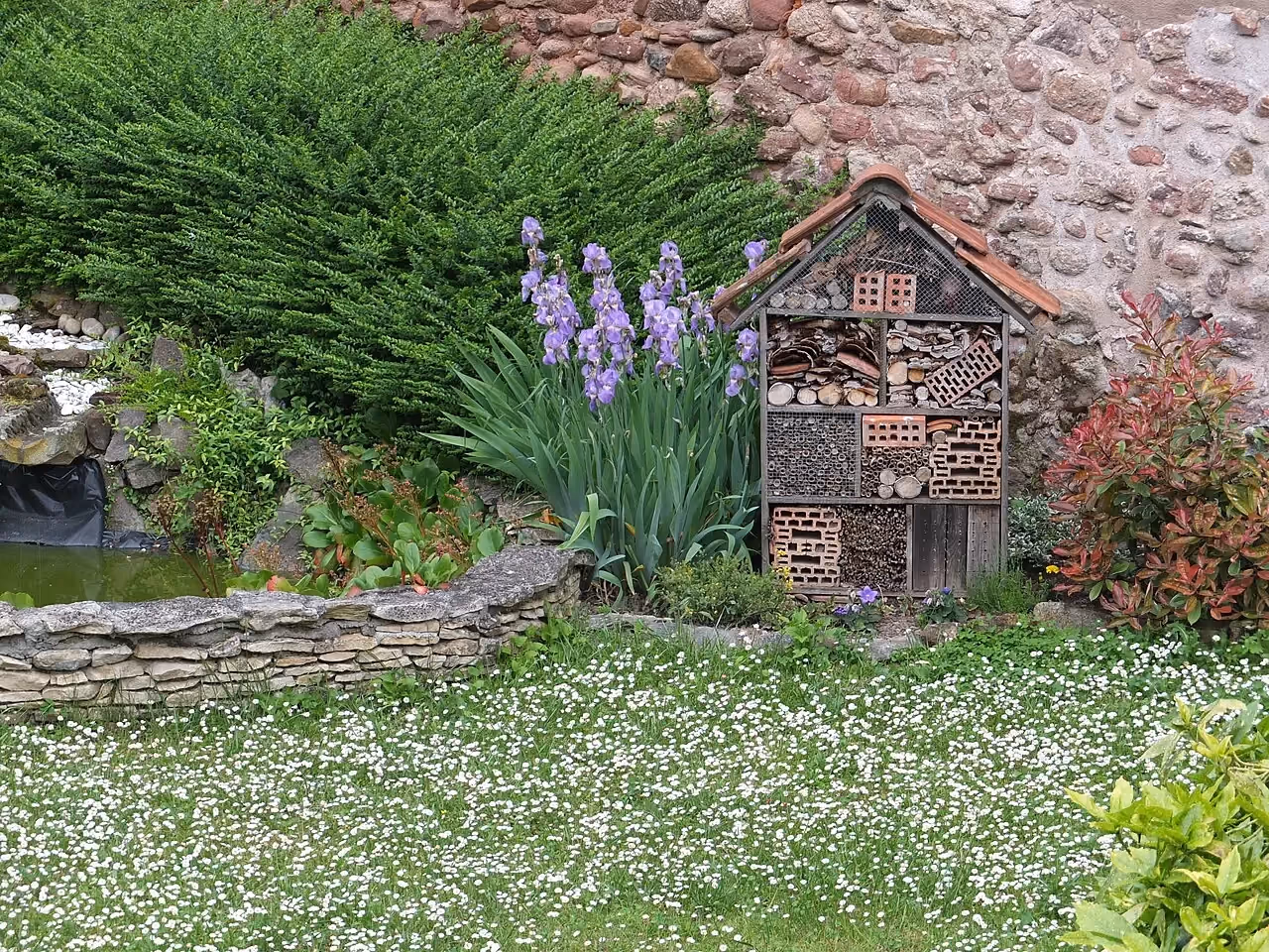 A wooden insect hotel with several compartments (hollow stems, drilled logs, bricks) in a garden in flower.