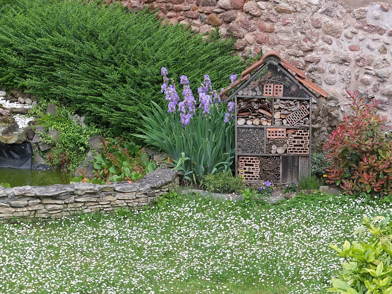 Un hôtel à insectes en bois à plusieurs compartiments (tiges creuses, bûches percées, briques) dans un jardin fleuri.